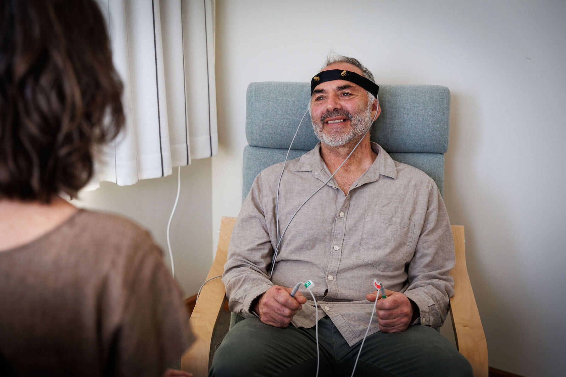 A smiling client relaxes in a chair during a bioresonance session, wearing a headband sensor and holding hand electrodes, with Caroline seated in the foreground